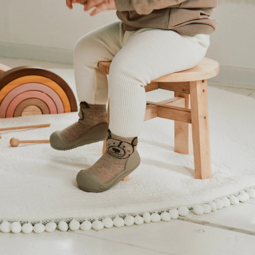 Niño pequeño sentado con zapatillas respetuosas bebe Attipas Teddy Beige, diseño de osito en tonos marrones