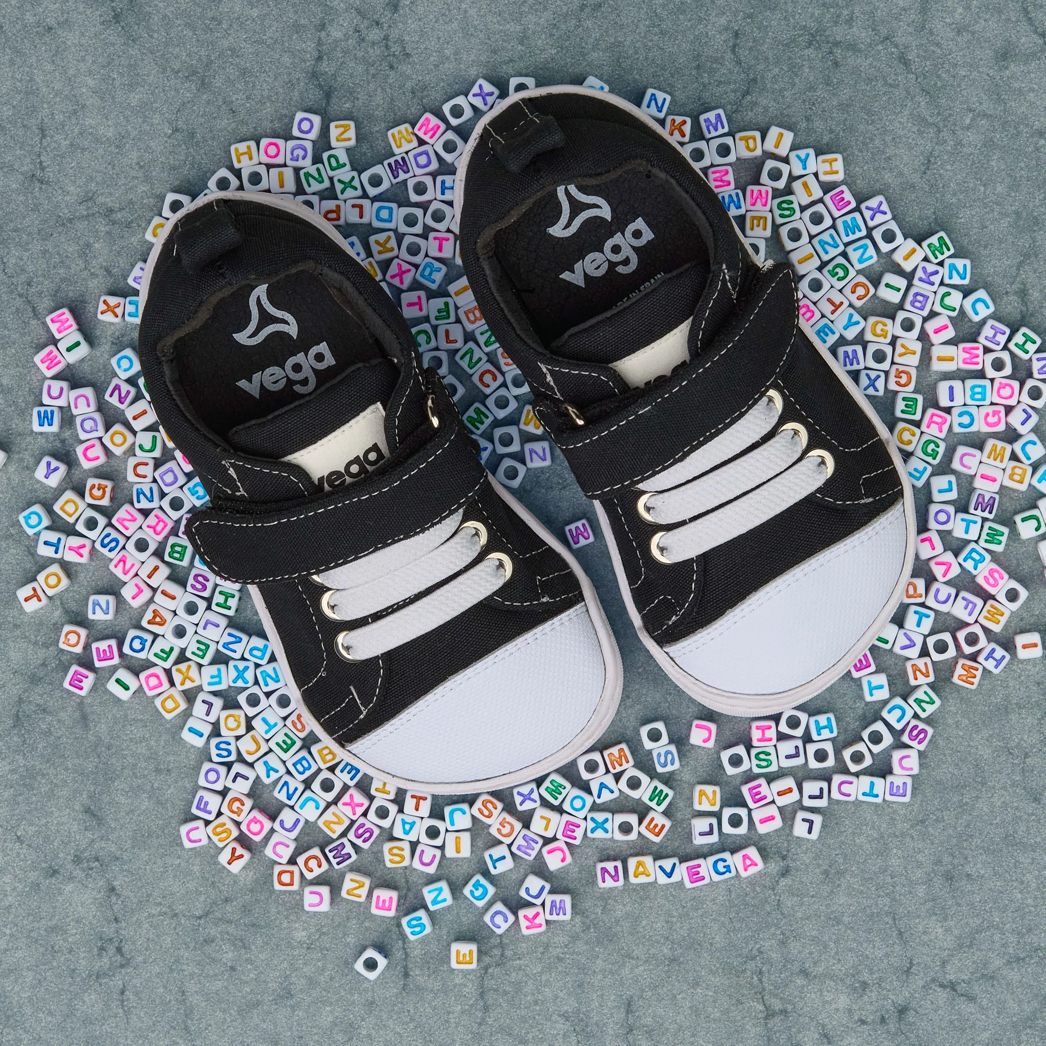 Zapatillas infantiles Vega Barefoot negras con puntera blanca, vista superior sobre fondo gris con abalorios de colores